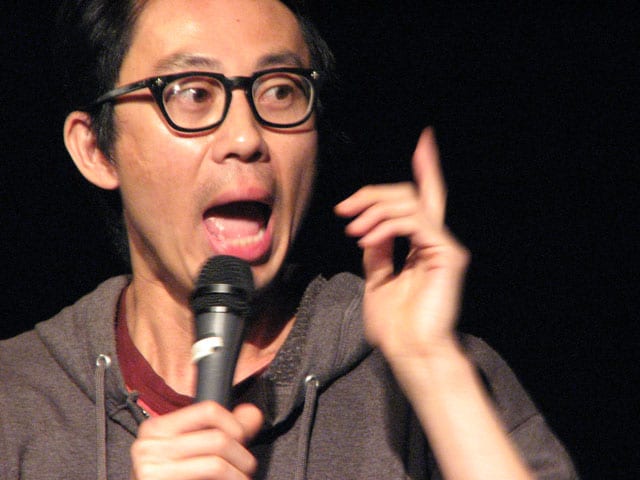 East asian man wearing black glasses with open mouth speaking into microphone