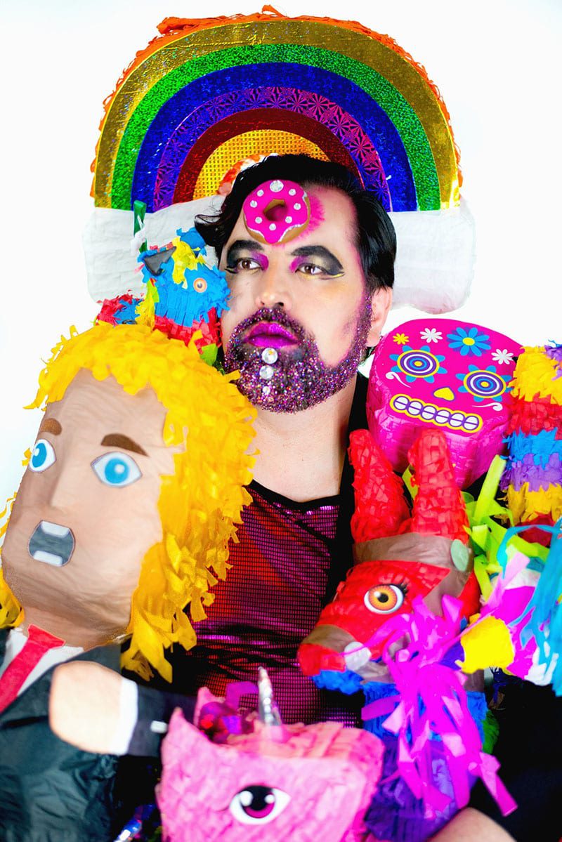 Person with dark hair and beard with pink lipstick surrounded by pinatas with a rainbow behind their head