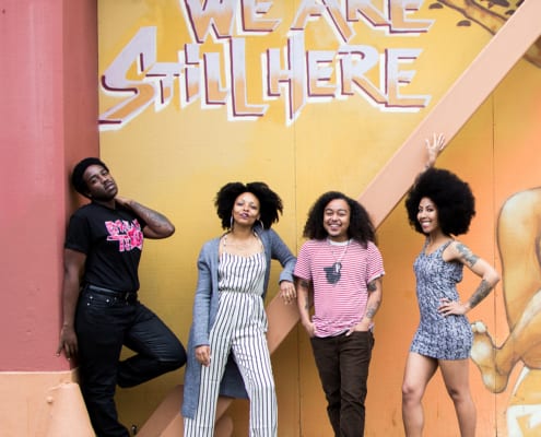 Group of black and brown people standing in front of a grafitti that says we are still here