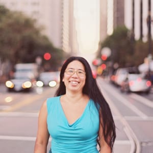 Long haired smiling person with glasses standing on a city street