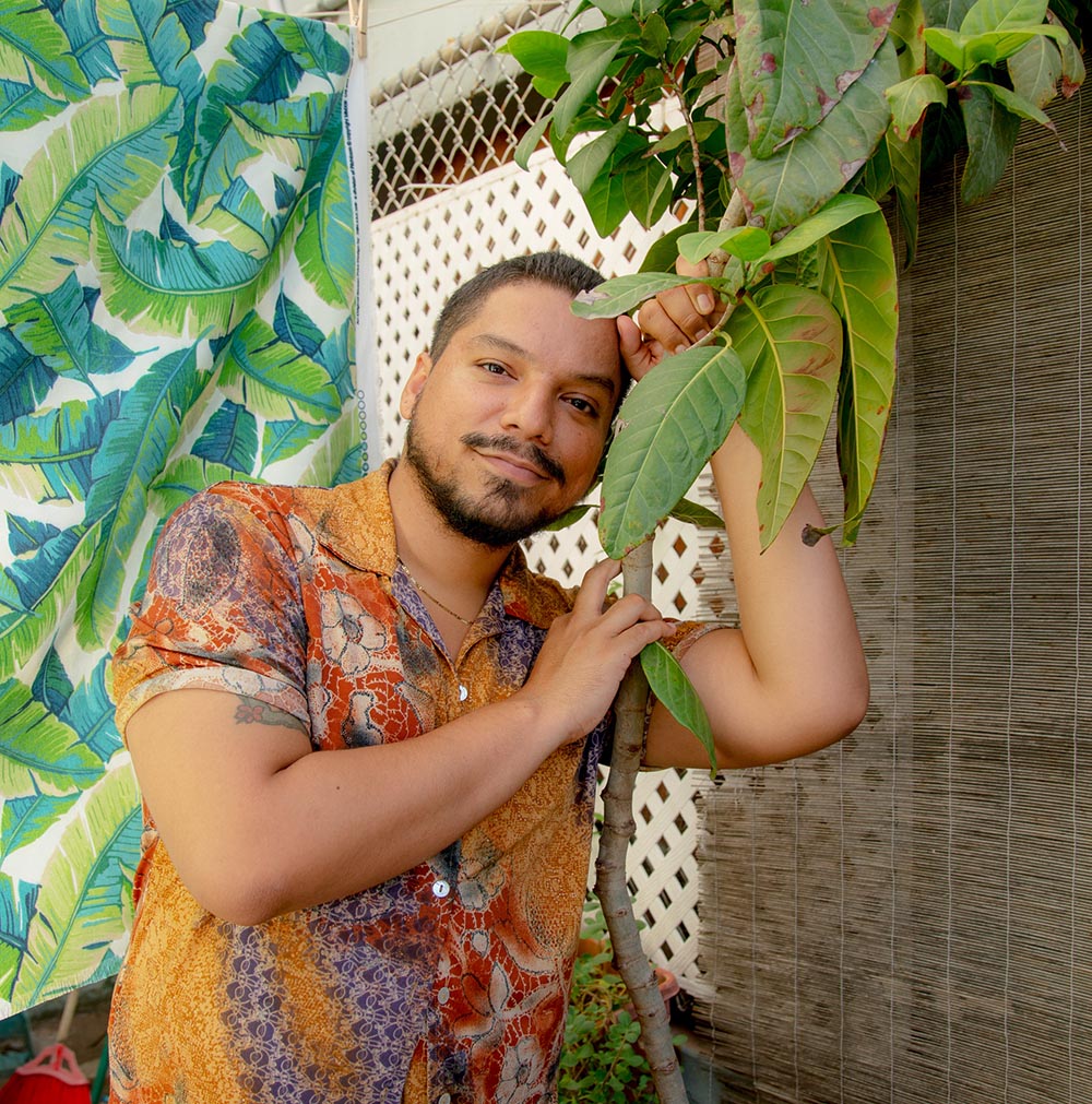 Brown skinned person leaning against a plant with short dark hair, beard, and mustache looking thoughtfully at the camera