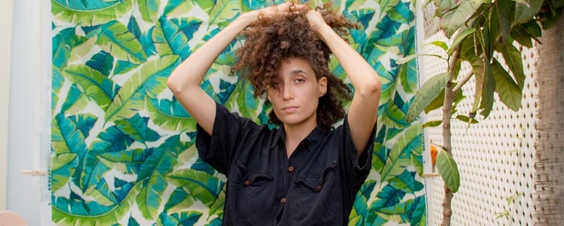 Brown curly haired person holding their hair up in black short sleeved jumpsuit standing against backdrop of green leaves in an outdoor space